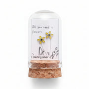 Small glass bottle with cork lid containing flowers, held over a cardboard box with floral elements.