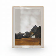 A framed landscape art print featuring a small white house on a golden-brown field with dark rocky mountains under a cloudy sky, set in a light wood frame against a white background.