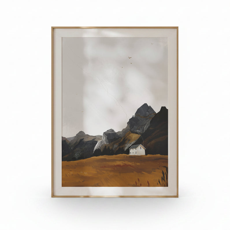 A framed landscape art print featuring a small white house on a golden-brown field with dark rocky mountains under a cloudy sky, set in a light wood frame against a white background.