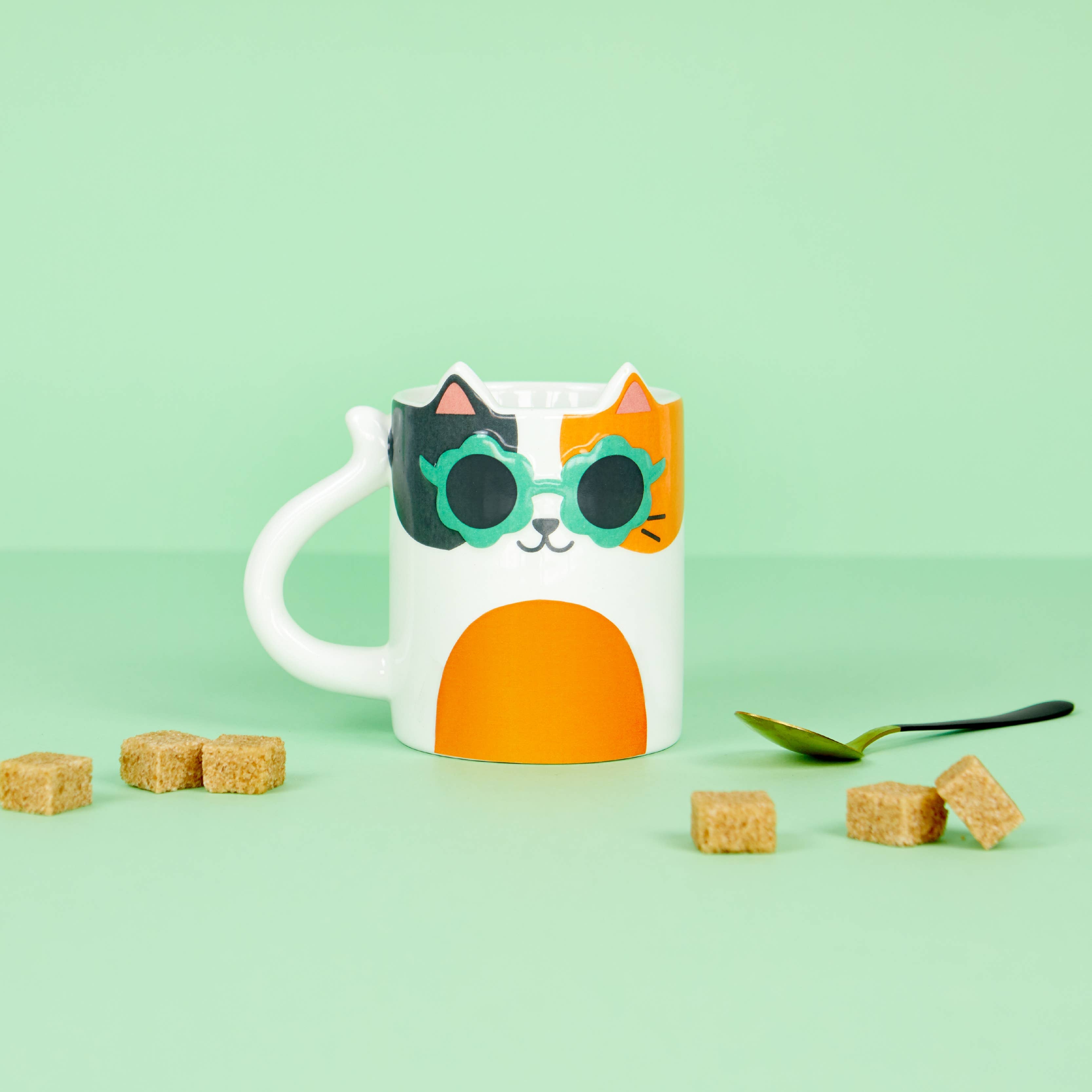 Cool Cat mug styled on a green background with sugar cubes and a spoon