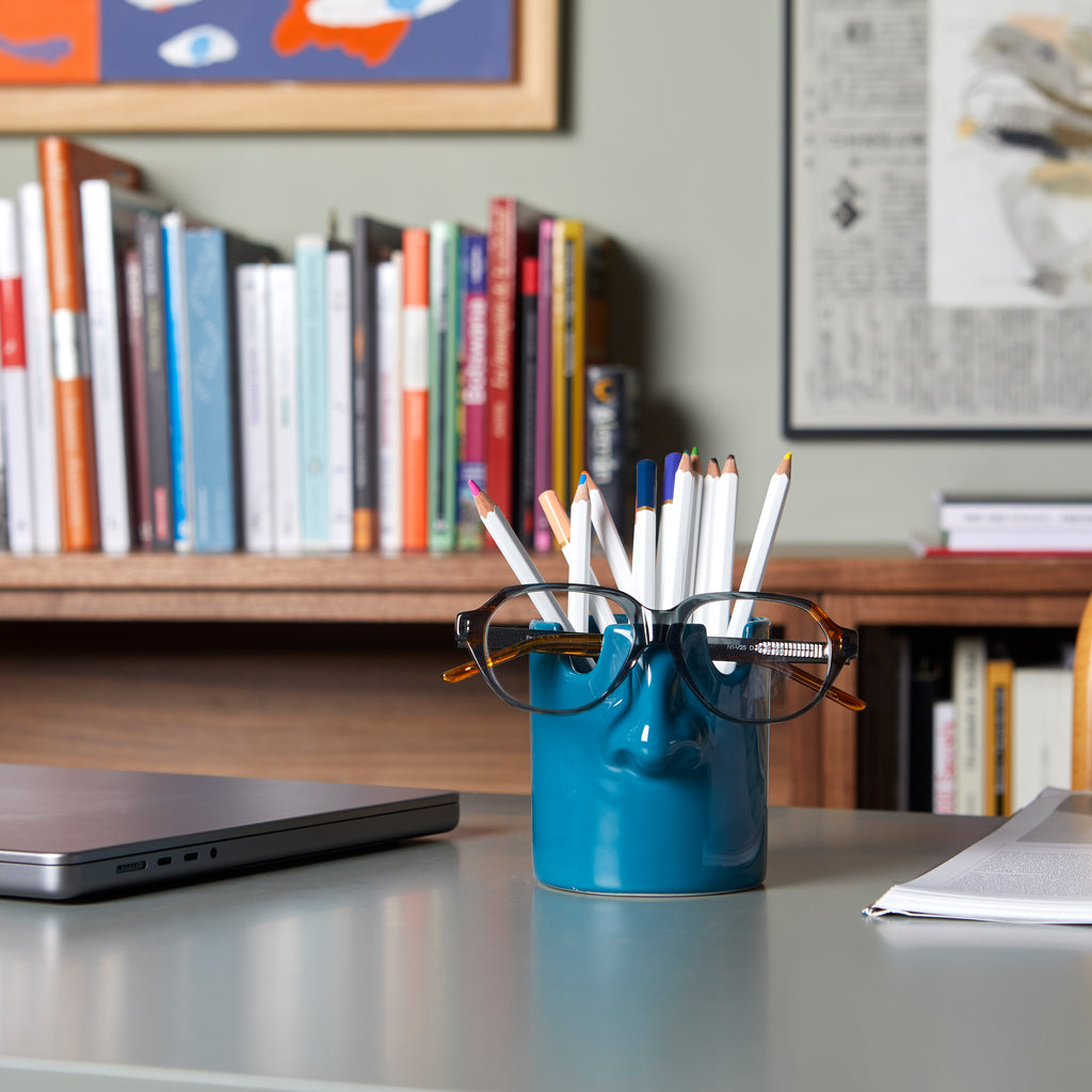 Teal nose pen holder on a desk in front of a bookshelf and laptop