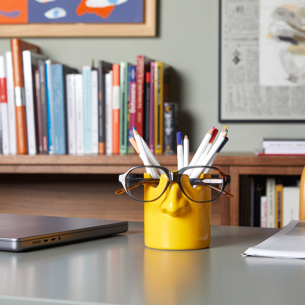 Mustard nose pen holder on a desk in front of a bookshelf and laptop