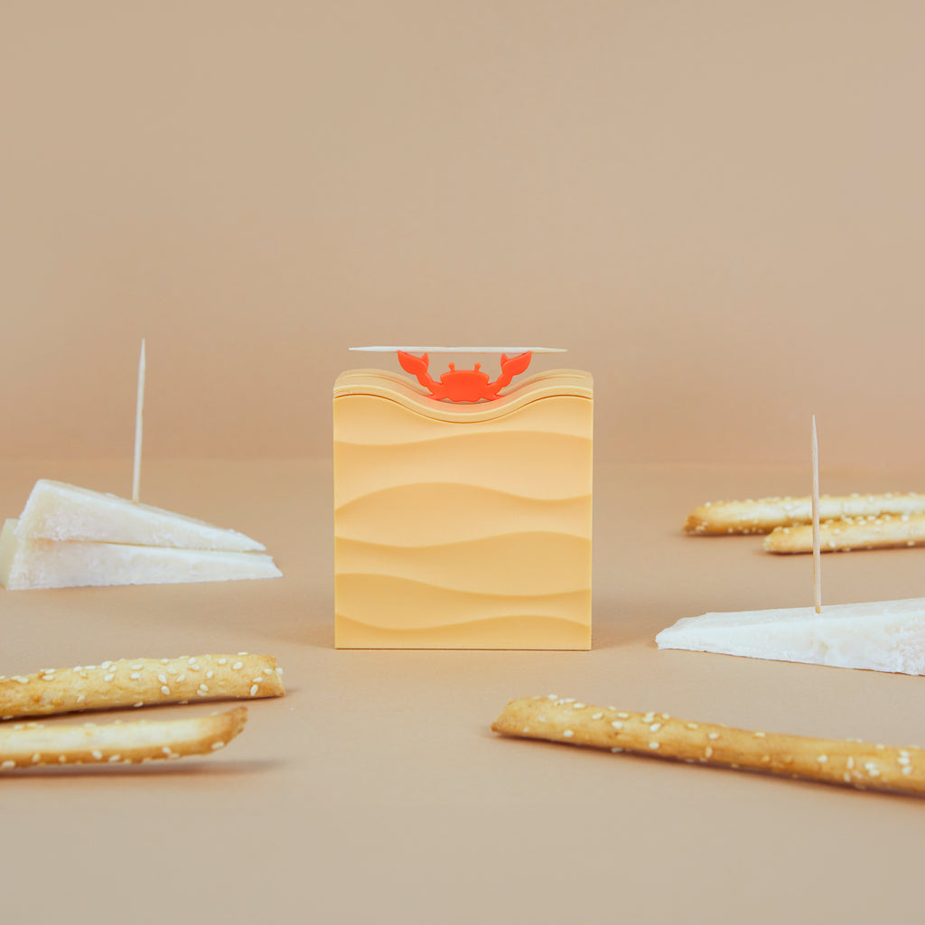 Crab toothpick dispenser on a beige table with crackers, brie and breadsticks