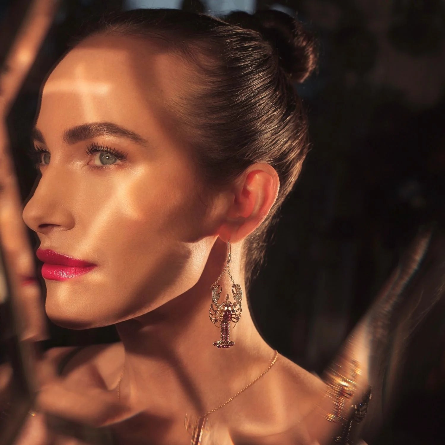 Close-up of woman wearing gold and red crystal lobster drop earring by Bill Skinner in warm lighting.