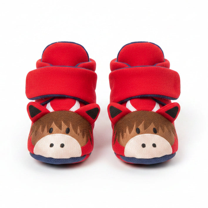 A close-up of a child's feet wearing red slipper-on slippers shaped like highland cows, paired with gray and navy striped tights.