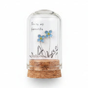 Small glass bottle with cork lid held by a hand, featuring a label with floral design and text on a wooden surface.
