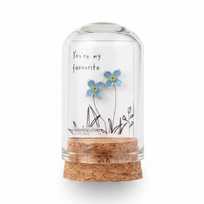 Small glass bottle with cork lid held by a hand, featuring a label with floral design and text on a wooden surface.