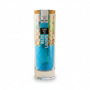 A tall clear glass jar with a cork stopper and printed label reading 'BLUE LAGOON' stands beside a tall clear glass filled with bright blue cocktail and a lemon slice, set on a sandy beach with the ocean and palm trees in the background.