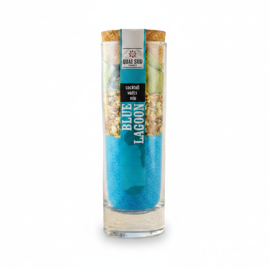 A tall clear glass jar with a cork stopper and printed label reading 'BLUE LAGOON' stands beside a tall clear glass filled with bright blue cocktail and a lemon slice, set on a sandy beach with the ocean and palm trees in the background.