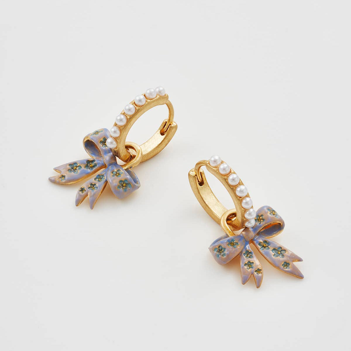 Pair of gold huggie earrings with pearl detail and hand-painted bow charms on a white background.