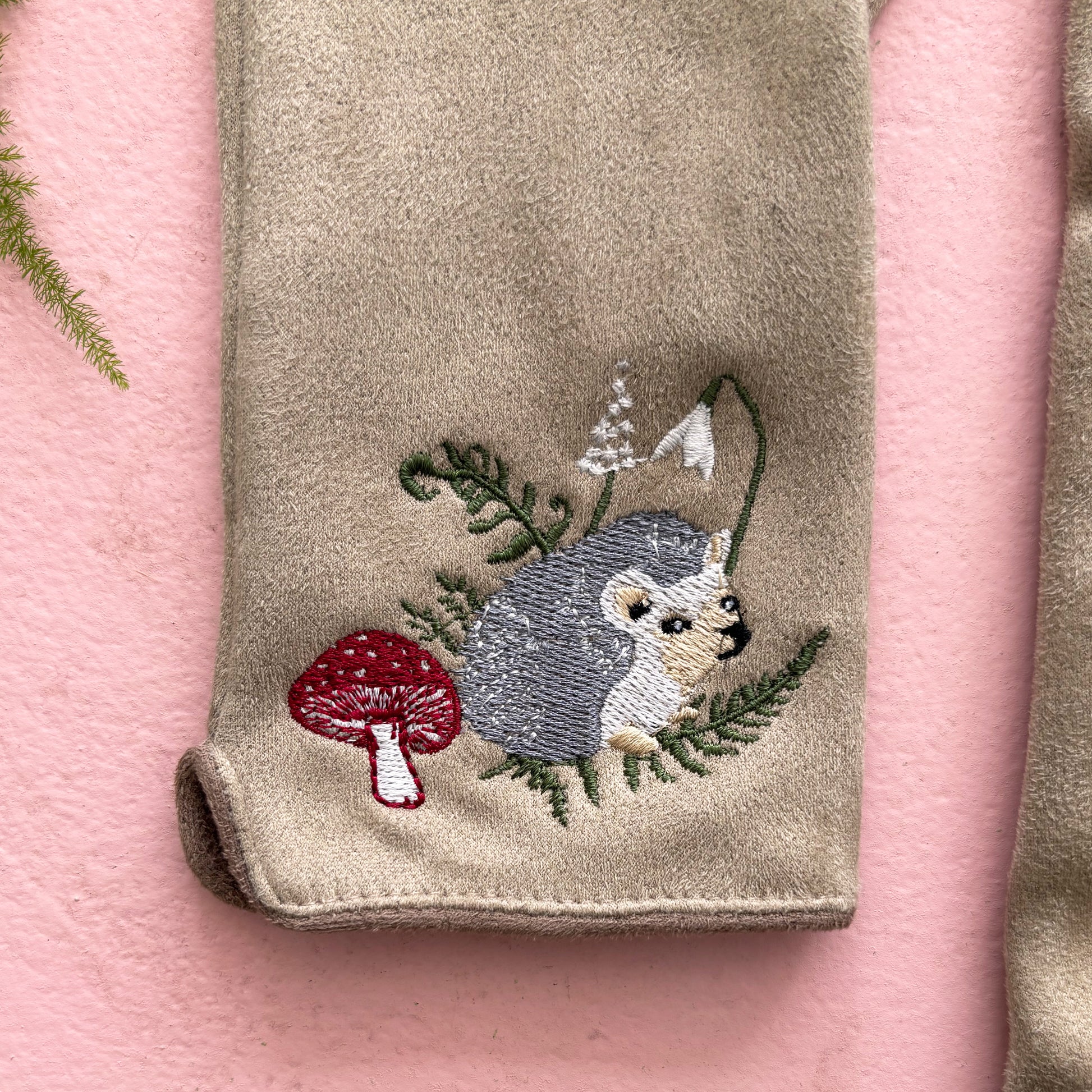 Detail of embroidered hedgehog among leaves and red mushroom on pink background