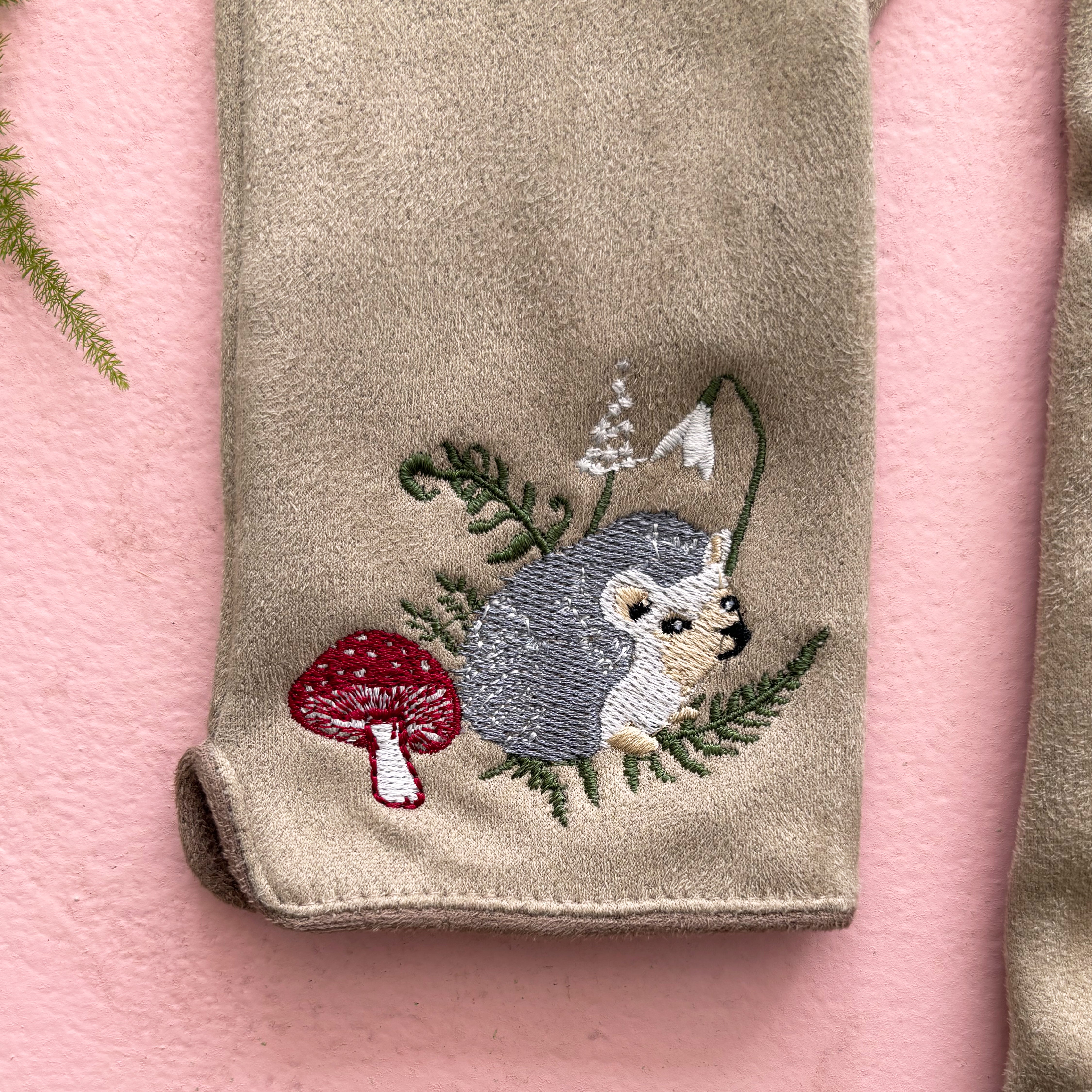 Detail of embroidered hedgehog among leaves and red mushroom on pink background