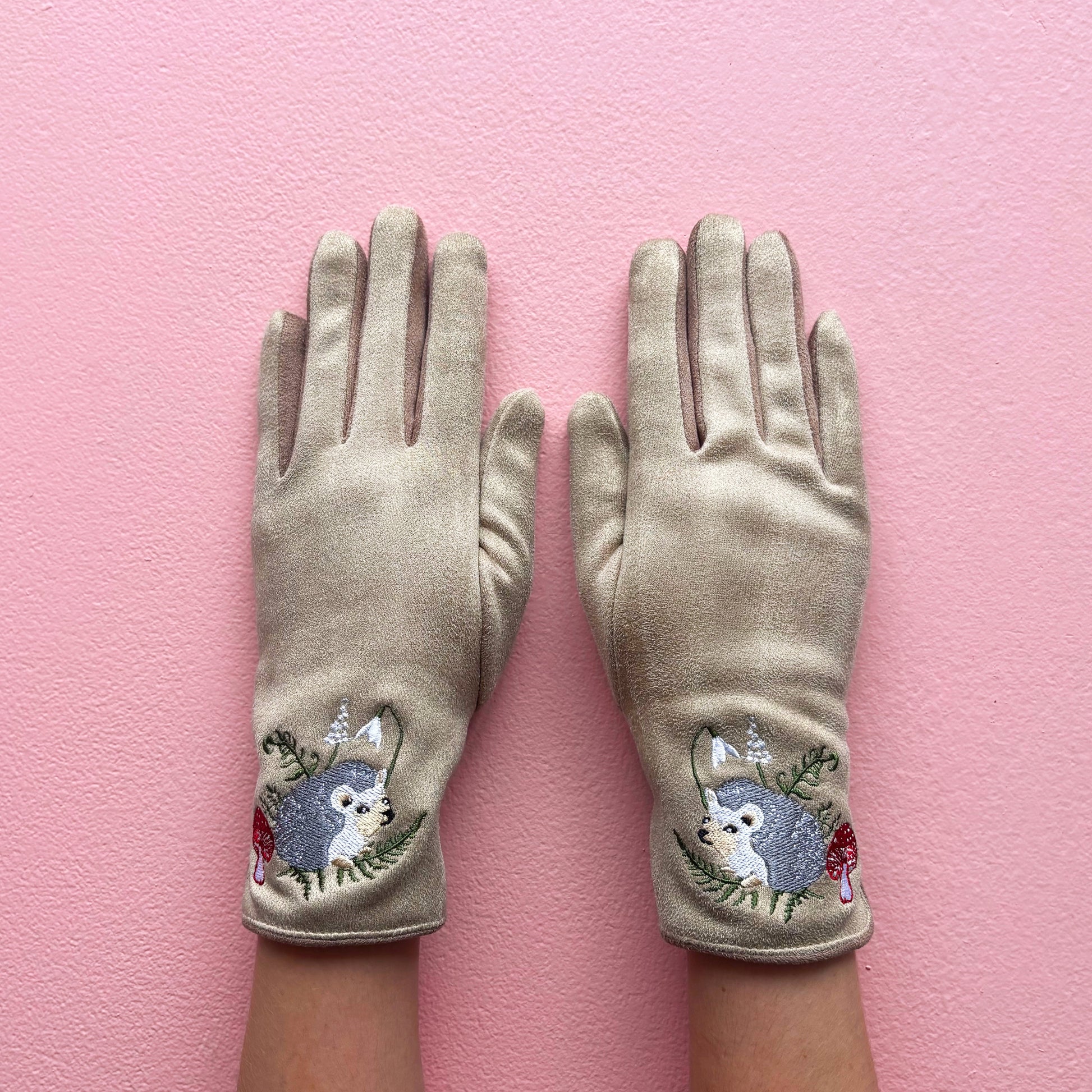 Beige embroidered hedgehog gloves worn on hands against pink background