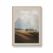A framed landscape art print featuring a misty mountain range, a lone cabin, and rolling hills in muted earth tones, displayed against a clean white background.
