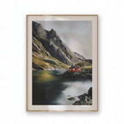 A framed landscape art print depicting a red cabin on a rocky shore next to a calm lake, surrounded by steep mountains and a misty sky, in a light wood frame with a white mat.