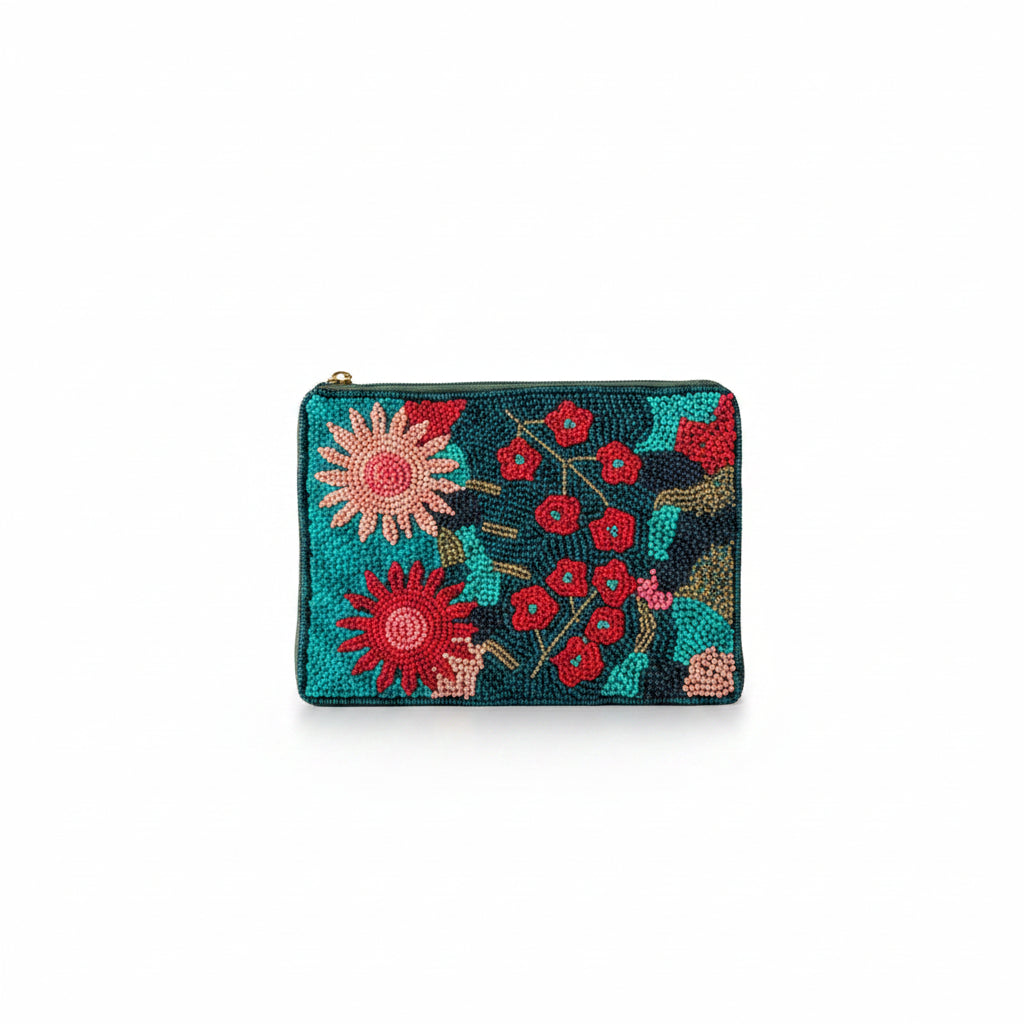 A person in a white dress holds a small zip-around clutch with a colorful floral needlepoint design. The clutch features a teal background with red, pink, and yellow flowers, set against a softly blurred indoor background with a lamp and chair.