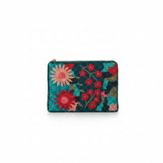 A person in a white dress holds a small zip-around clutch with a colorful floral needlepoint design. The clutch features a teal background with red, pink, and yellow flowers, set against a softly blurred indoor background with a lamp and chair.