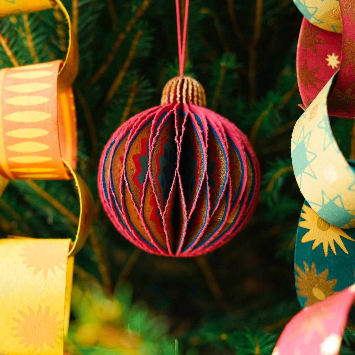 Pink Bauble Honeycomb Christmas decoration with gold cap hanging on a fir tree beside paper chains