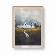 A framed landscape art print depicting a white cottage with a gray roof nestled in a grassy valley with mountains in the background, enclosed in a light wood frame.