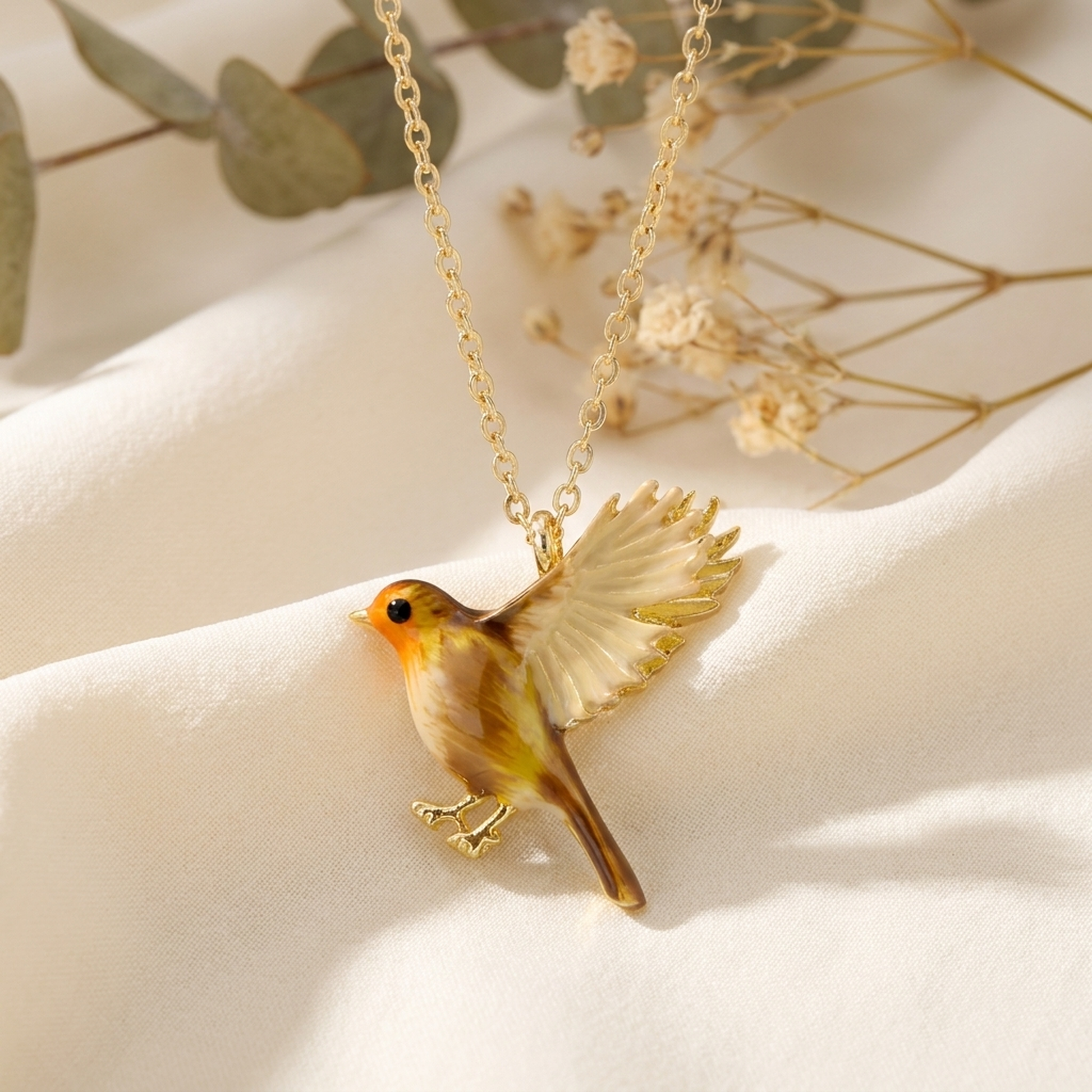 Enamel Flying Robin Necklace draped on soft cream fabric with dried flowers