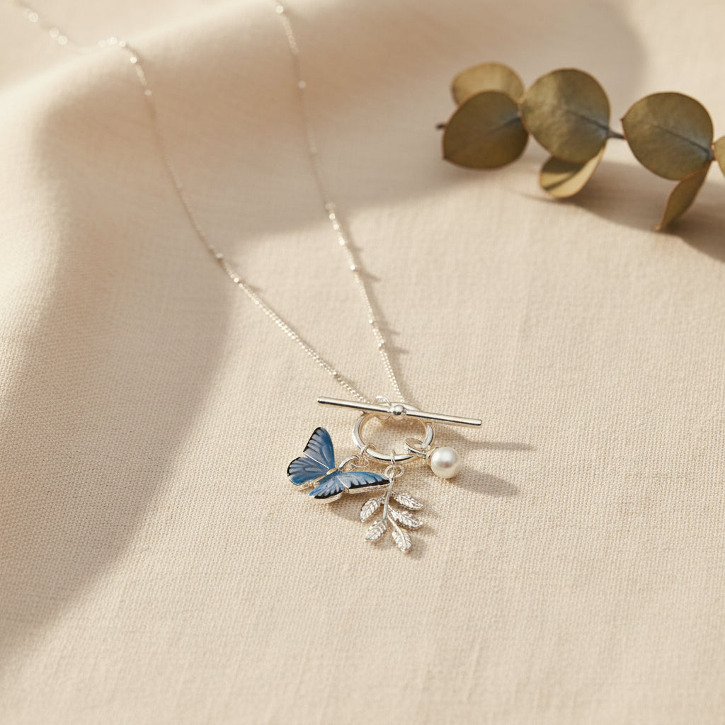 A silver toggle clasp necklace featuring a butterfly charm with blue and black wings, a silver branch with leaves, and a small pearl bead, shown on a light gray background.