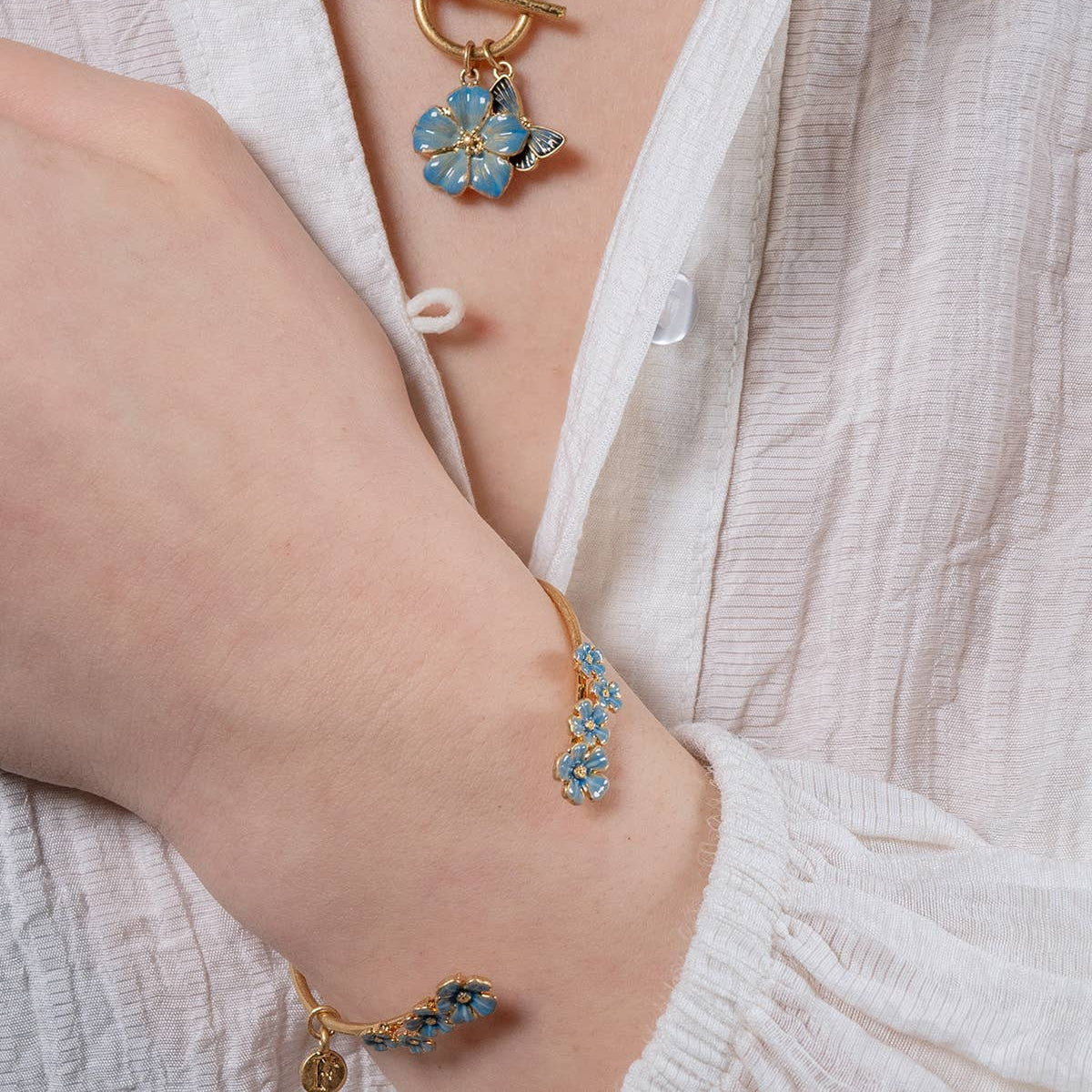 Gold-plated forget-me-not bangle worn on wrist with matching blue enamel flower necklace