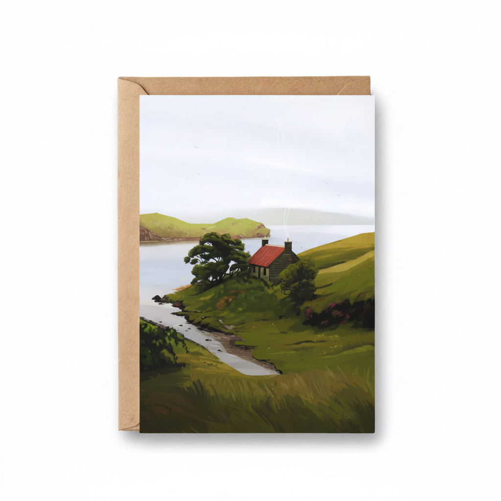 A vertical art print or postcard featuring a painted coastal landscape with green hills, a small house with a red roof, trees, and calm water, shown standing against a kraft paper envelope on a light wood surface.