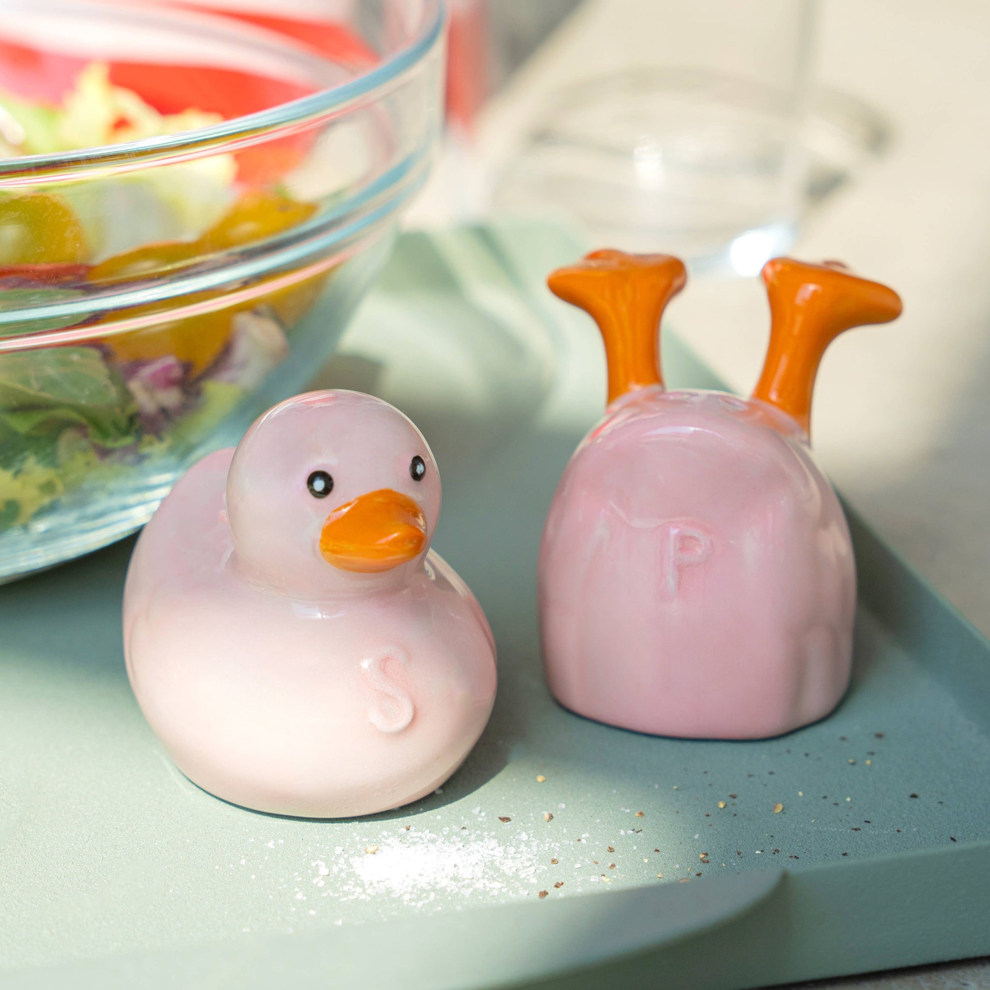 Pink duck salt and pepper shakers on a green board beside a salad bowl