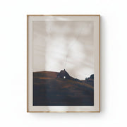 A framed landscape art print featuring a small cottage on a hill under a cloudy sky, displayed on a clean white wall.
