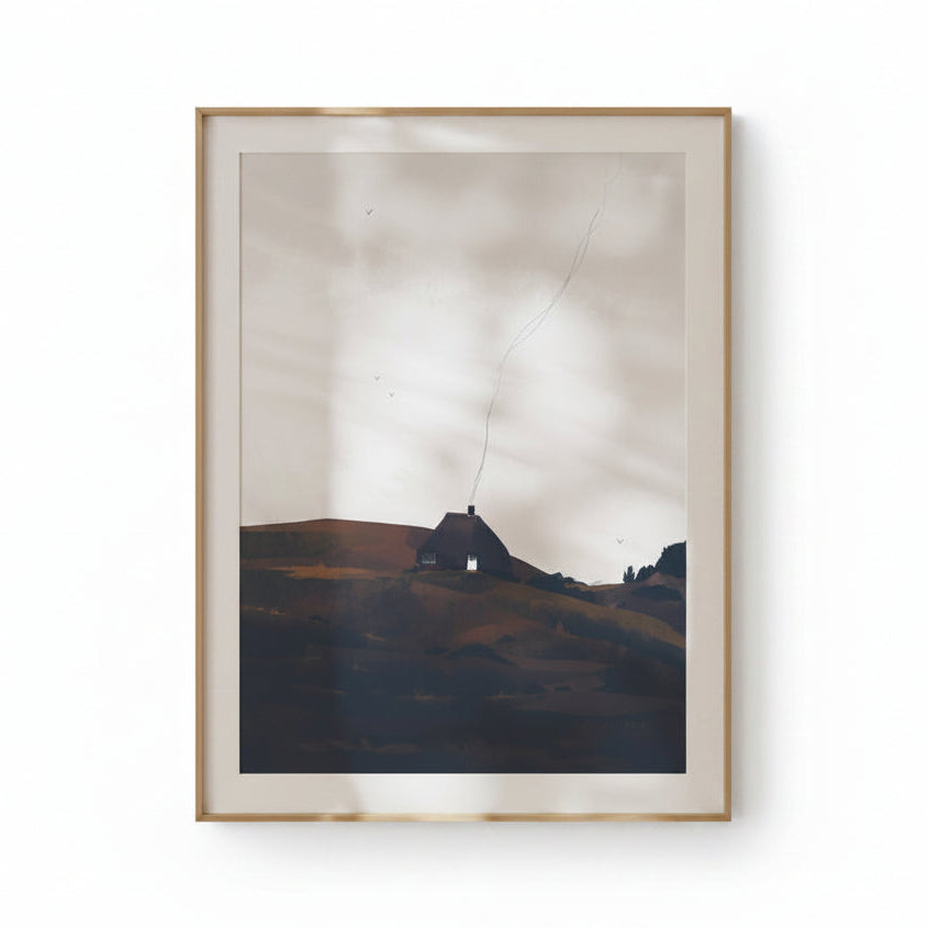 A framed landscape art print featuring a small cottage on a hill under a cloudy sky, displayed on a clean white wall.