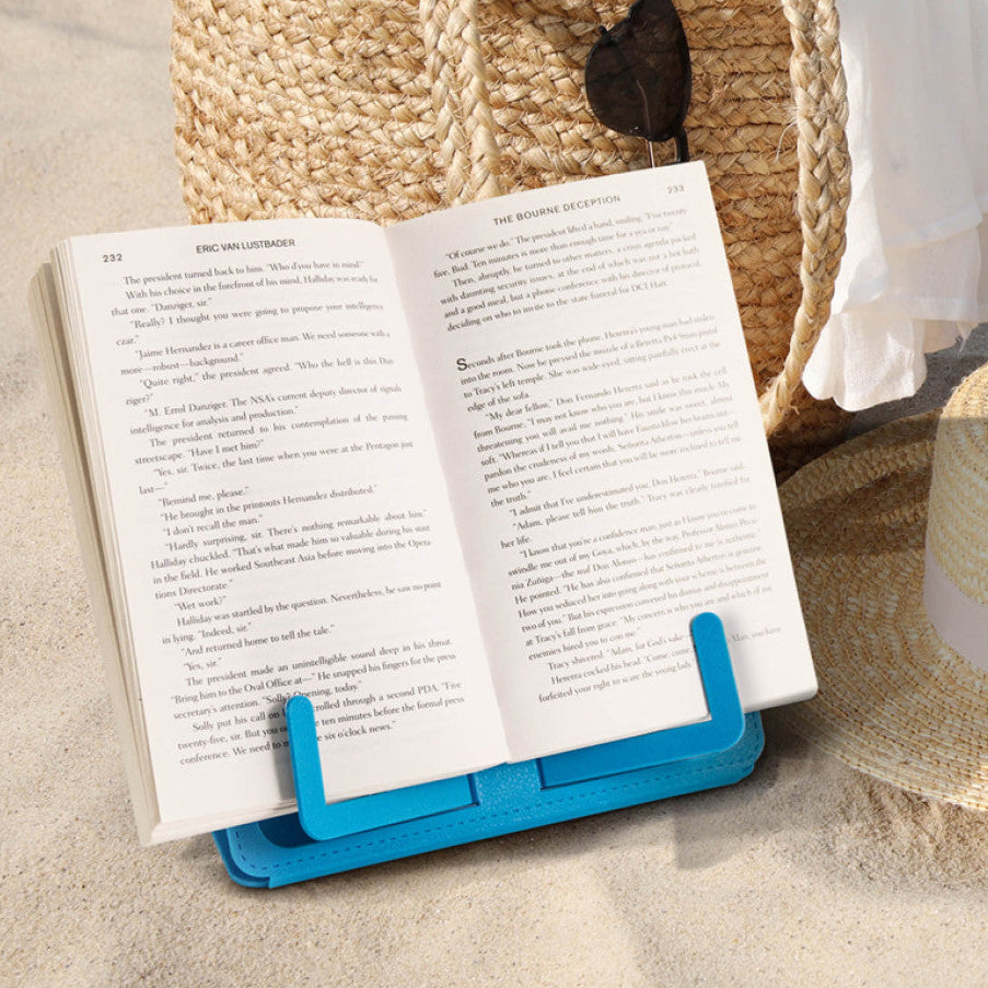 Blue Travel Book Rest holding an open paperback beside a straw beach bag