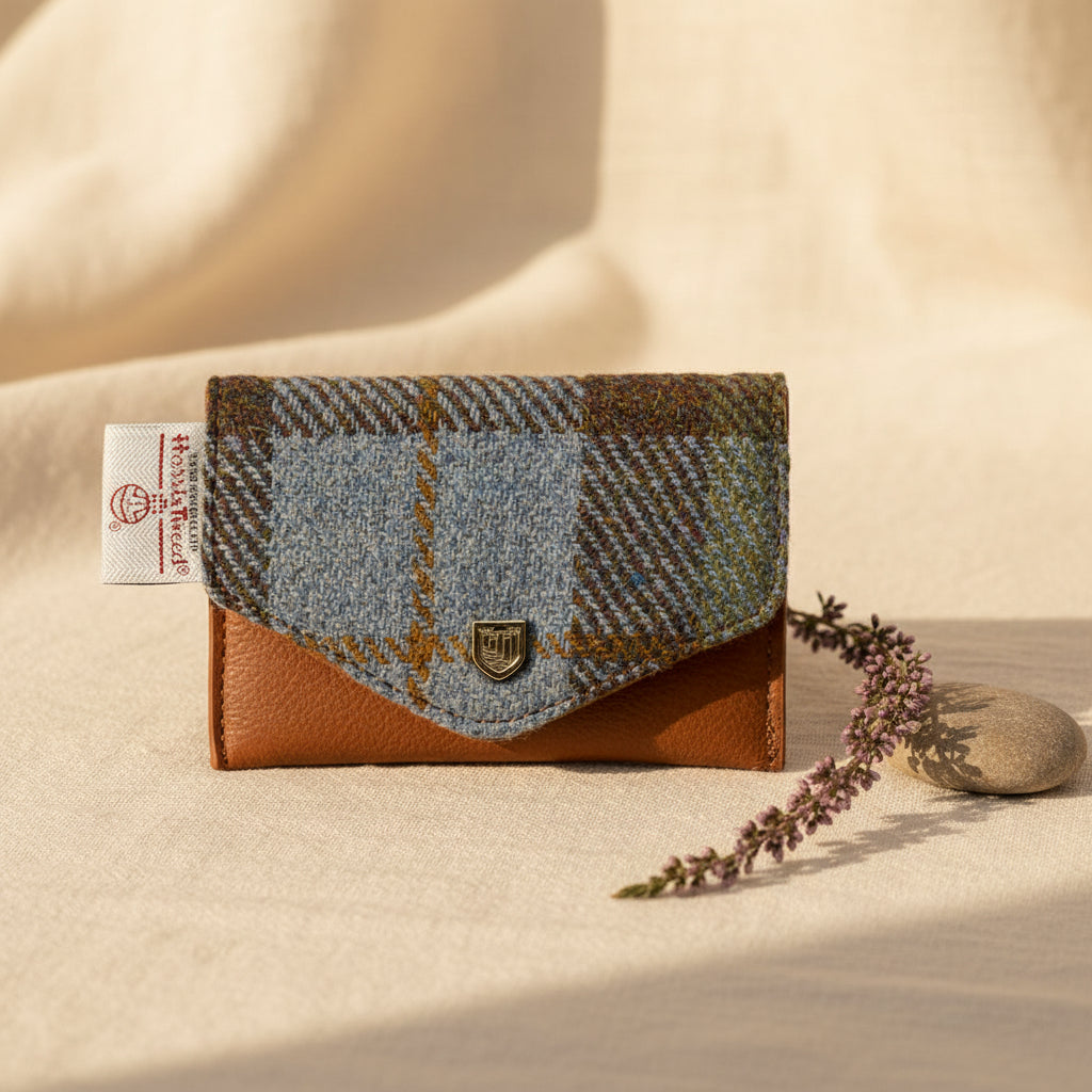 A small tweed wallet with a brown leather base and flap, featuring a blue and brown plaid pattern, a white fabric brand tag on the left, and a small gold crest emblem on the front.
