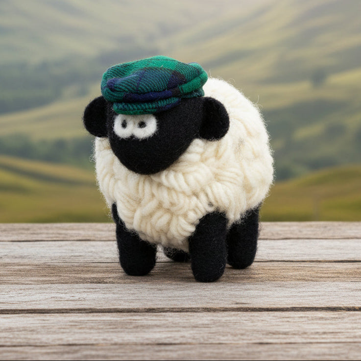 A small felt sheep toy with fluffy white wool, a black face and legs, and a green tartan cap, photographed on a plain white background.