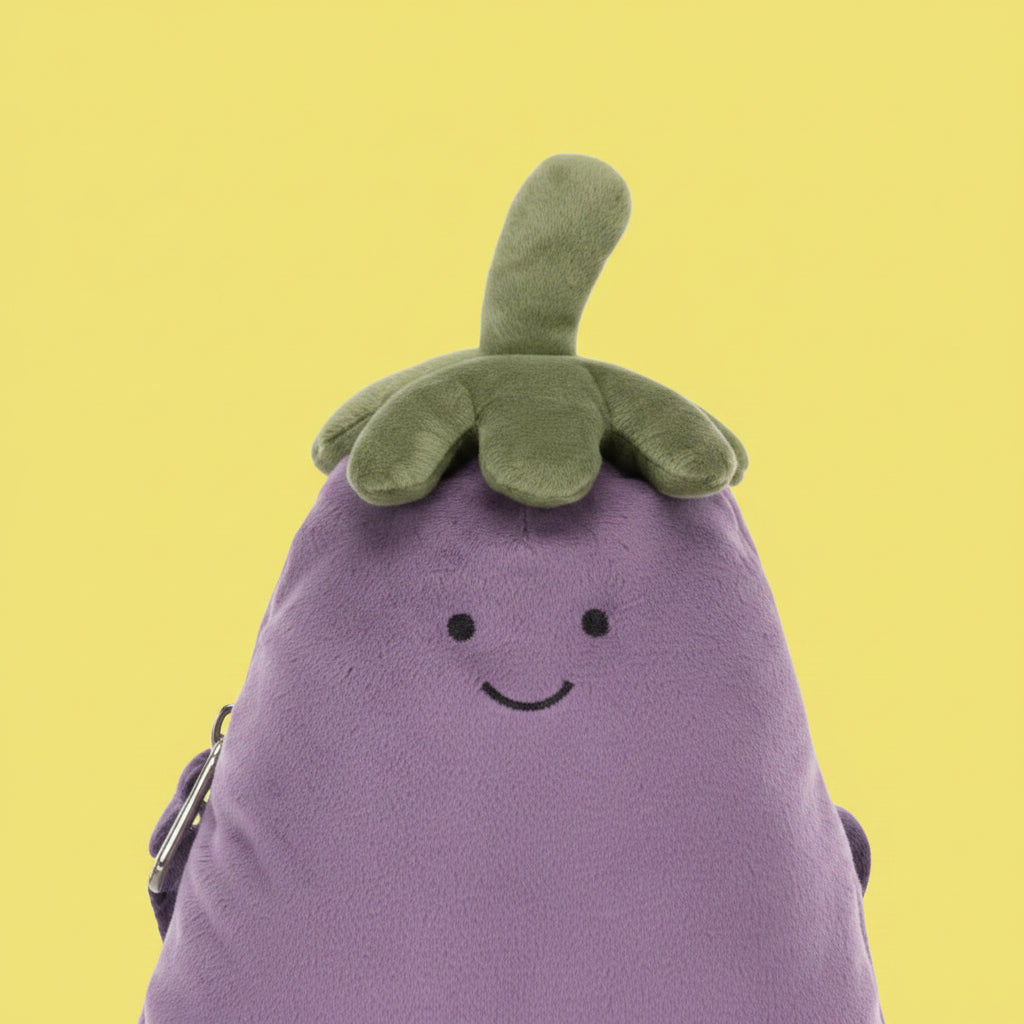 A purple egg-shaped crossbody bag with a green plush top and a smiling embroidered face, shown on a clean white background with its matching strap laid beside it.