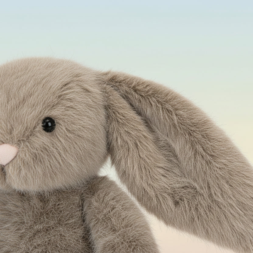 A plush stuffed bunny in a warm taupe-gray color with long floppy ears, a small pink nose, and black bead eyes, photographed on a clean white background.