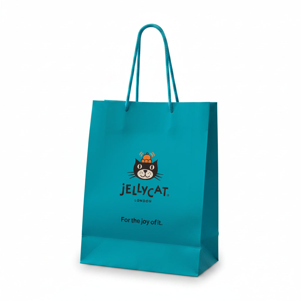 A hand holds a bright turquoise Jellycat paper shopping bag with the brand's cat logo and 'Jellycat London' text, set against a softly blurred indoor background with shelves and plush toys.