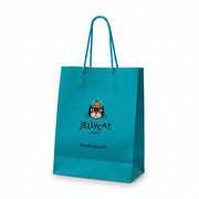 A hand holds a bright turquoise Jellycat paper shopping bag with the brand's cat logo and 'Jellycat London' text, set against a softly blurred indoor background with shelves and plush toys.