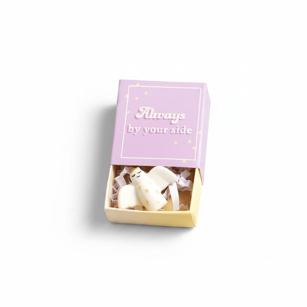 Ceramic angel nestled inside open lilac matchbox on cream background