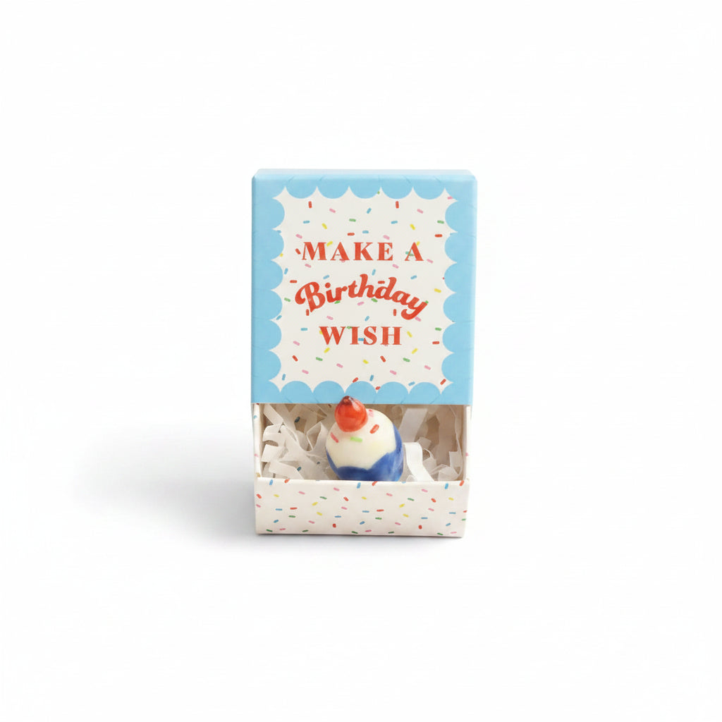 Ceramic cupcake inside open confetti matchbox with blue lid on cream background