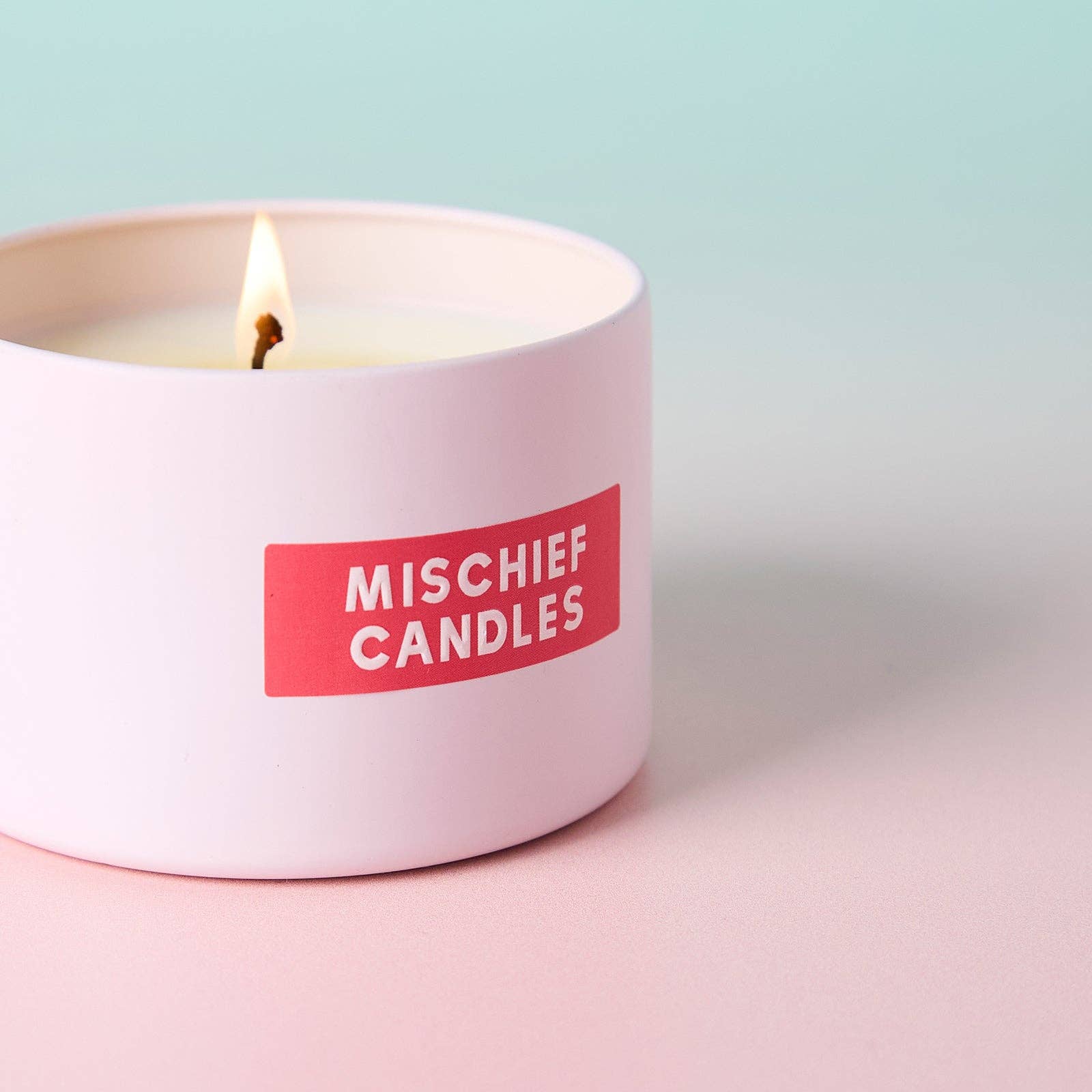 Mischief Candles pink tin close-up with lit wick on pastel background