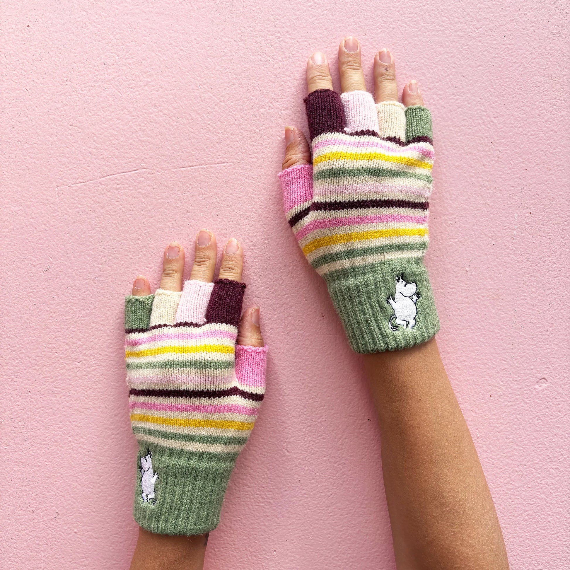 Hands wearing Moomin Stripy Fingerless Gloves with pastel and berry-toned stripes against a pink background.
