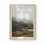 Framed landscape painting of a house on a hillside against a white wall.