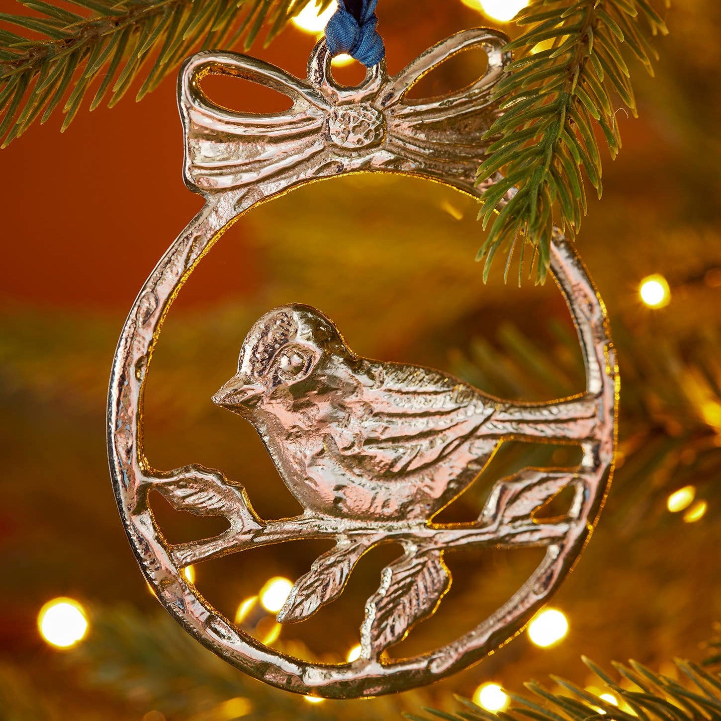 A silver metal Christmas ornament shaped like a bird perched on a branch inside an oval frame with a bow at the top, hanging from a blue ribbon against a blurred Christmas tree background with warm lights.
