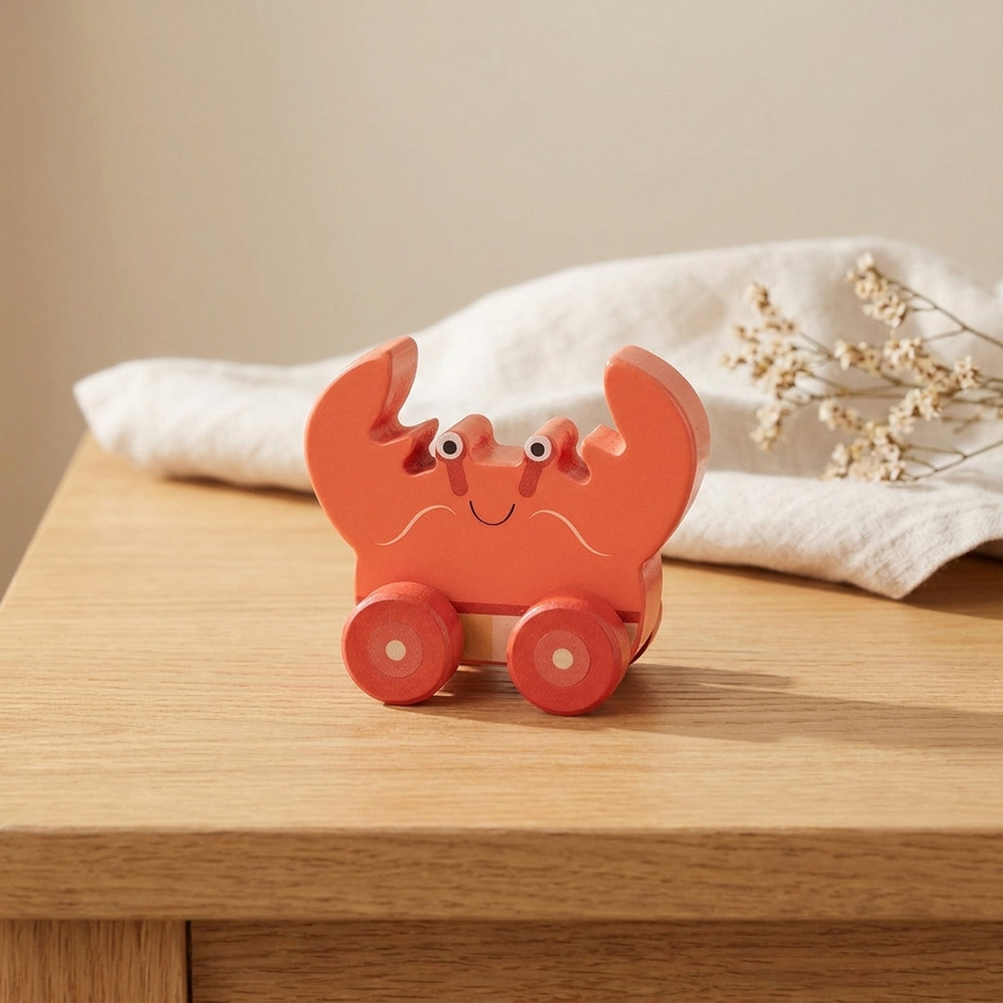 Orange wooden crab push toy on a warm oak table with cream linen and dried flowers