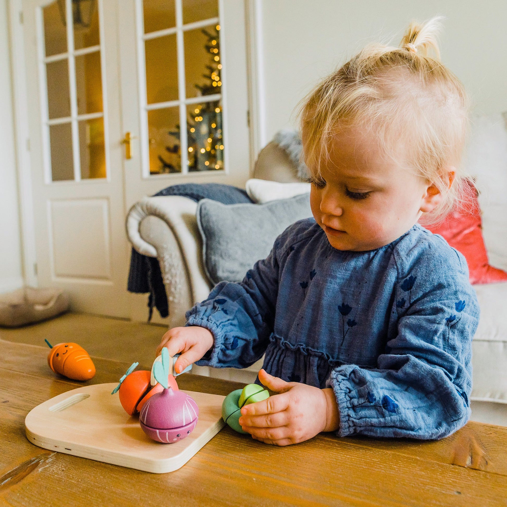 Toddler slicing a wooden carrot on the board at a wooden kitchen table