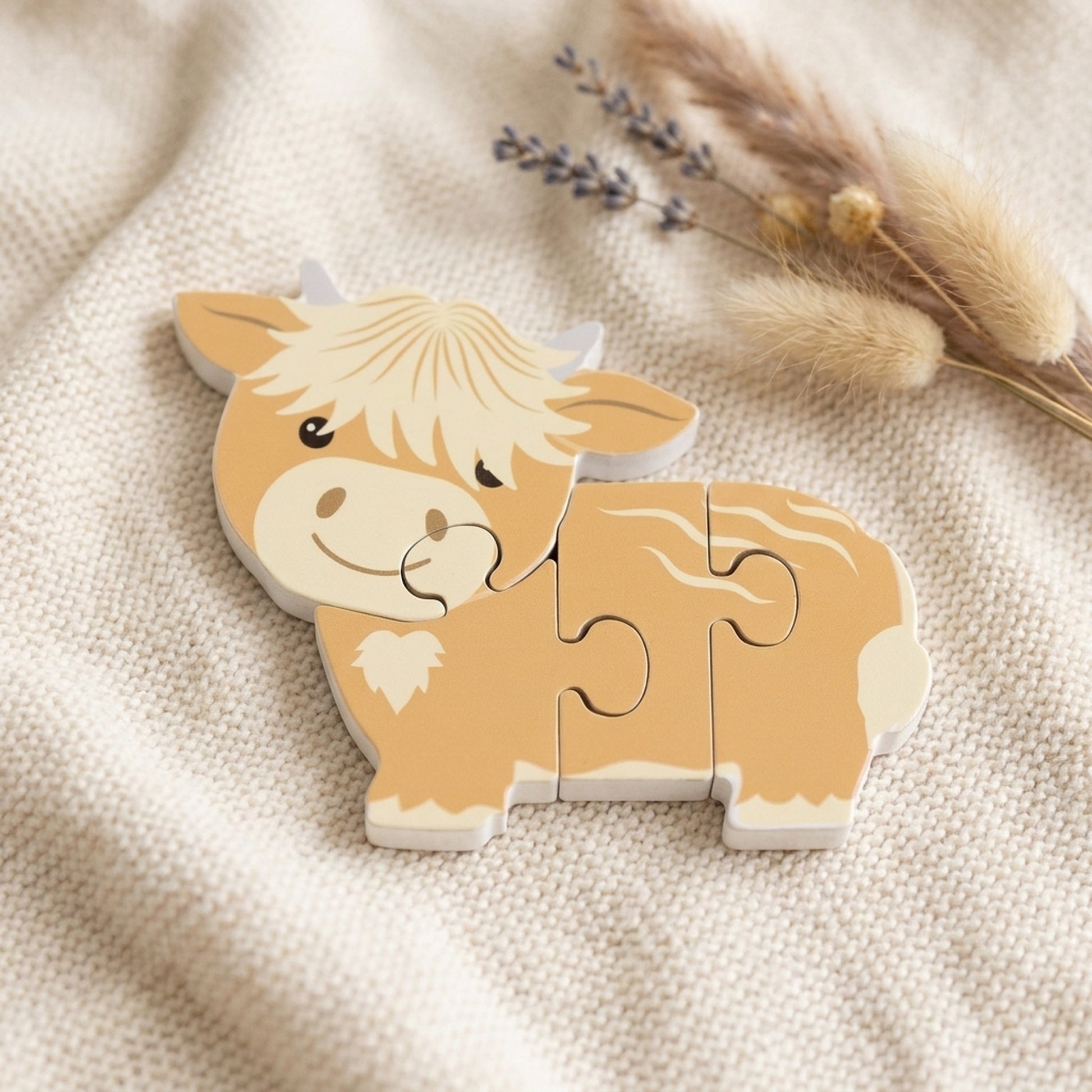 Highland cow wooden puzzle resting on a soft cream knitted blanket with dried flowers