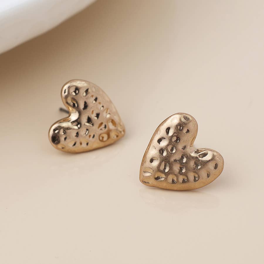 Gold hammered heart stud earrings beside a white dish on cream