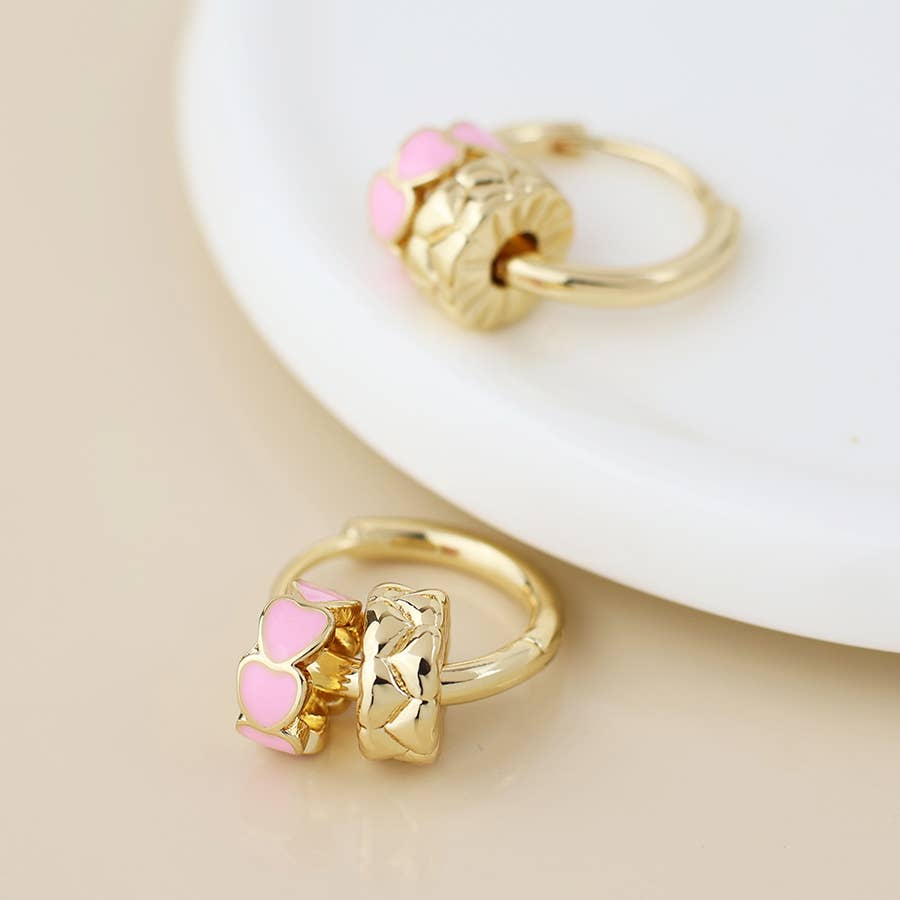 Gold hoop earrings with pink enamel and embossed heart rings on a cream surface