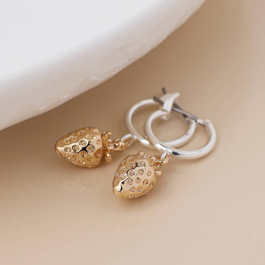 Gold strawberry charm silver hoops beside a white dish
