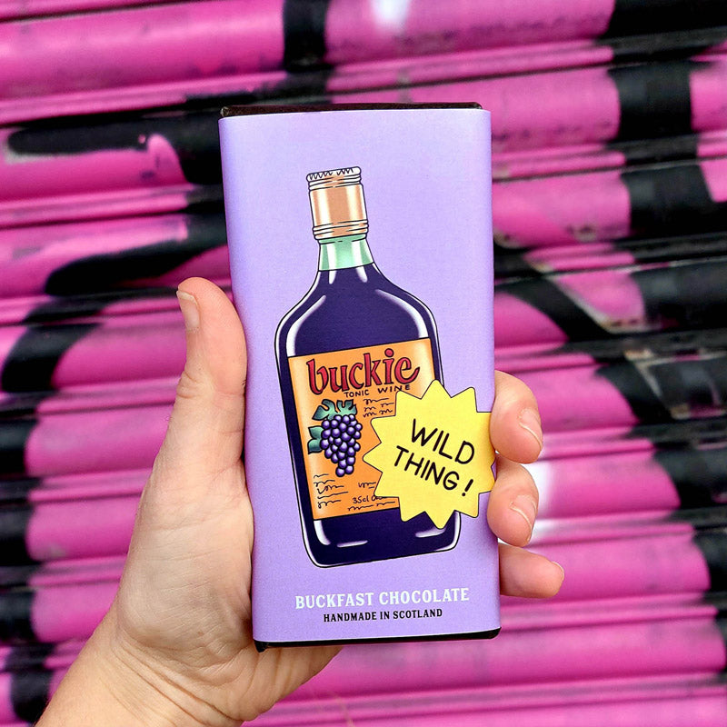 Buckfast chocolate bar in lilac buckie bottle wrapper held against pink graffiti